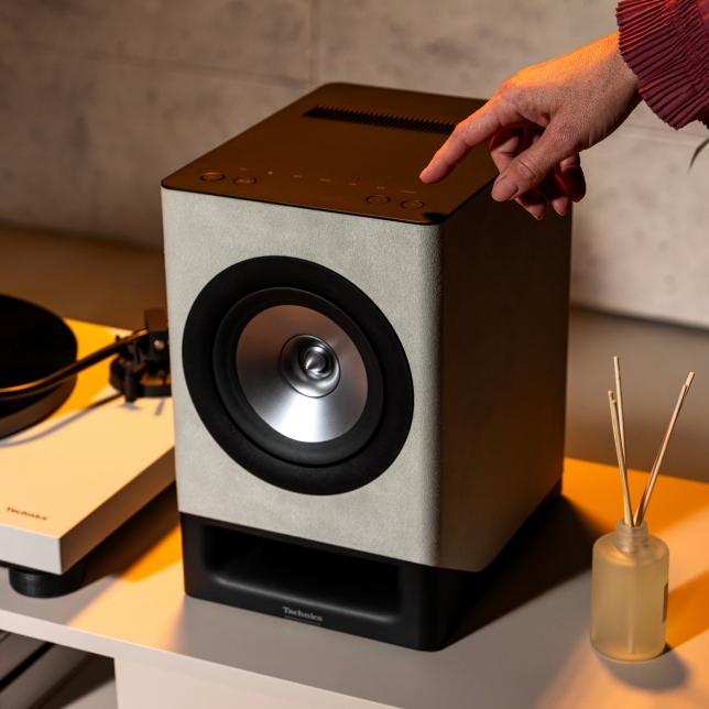 Details of the Technics SC-CX700 wireless Hi-Fi speaker in a stylish living room, in light grey colour. We can see a hand operating the button on top of the product.