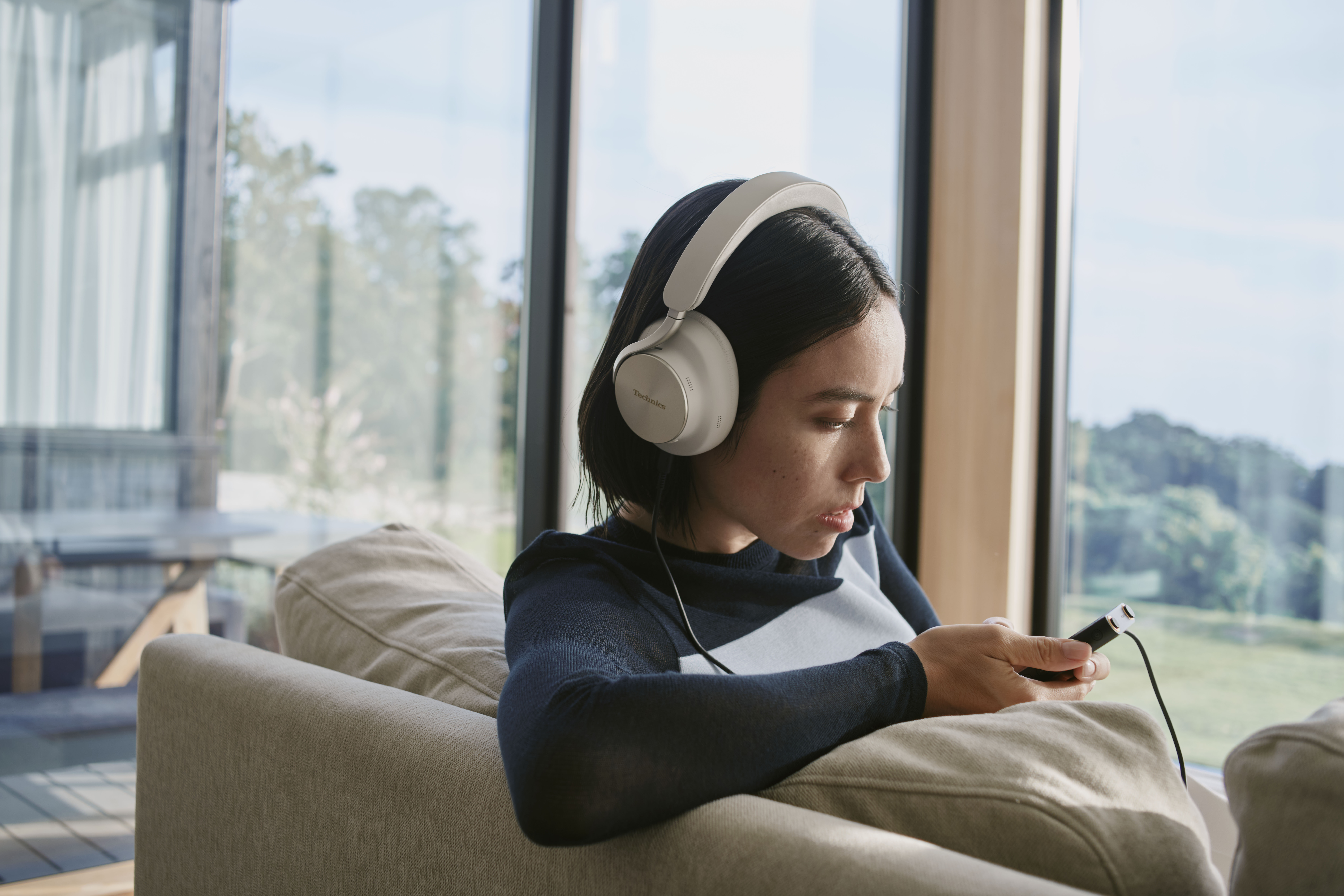 Una donna ascolta musica su un lettore audio portatile (DAP) tramite connessione cablata, indossando cuffie circumaurali Technics color grigio-beige.