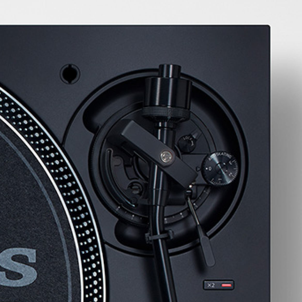 This image shows a close-up top view of a turntable tonearm base and control assembly, featuring a black S-shaped tonearm with adjustable settings and a pitch control dial beside a partially visible platter.