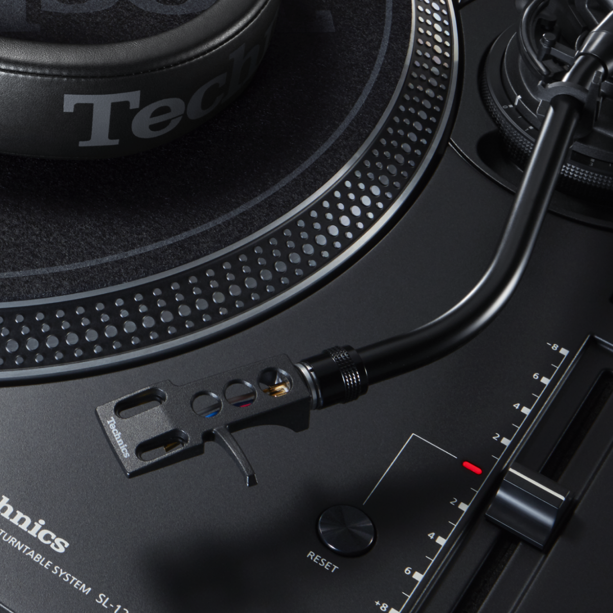 This image shows a close-up of a turntable’s S-shaped tonearm and cartridge positioned near the platter edge, alongside a vertical pitch control fader with scale markings and a reset button on a matte black deck.