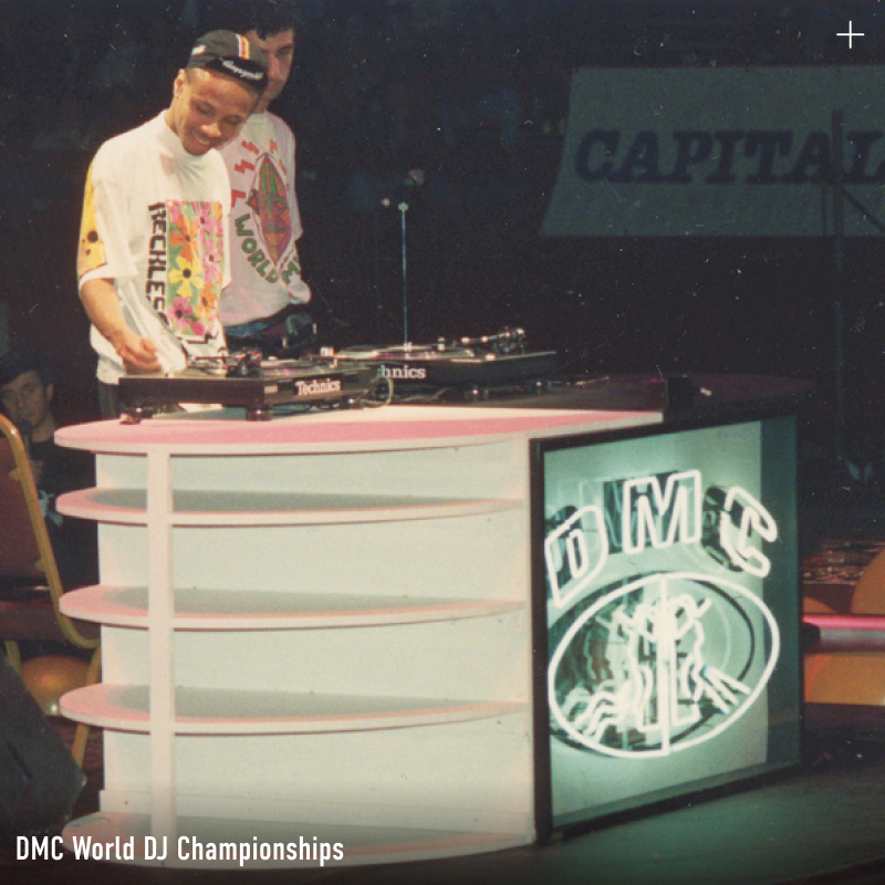 A vintage photograph of a DJ competing on Technics turntables at the DMC World DJ Championships, with the DMC logo displayed on a screen beside the stage.