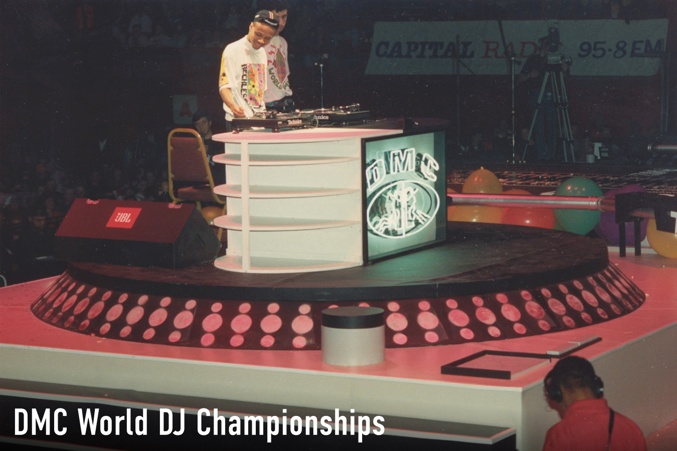 A vintage photograph of a DJ competing on Technics turntables at the DMC World DJ Championships, with the DMC logo displayed on a screen beside the stage.