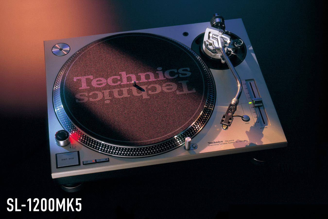 Photograph of the Technics SL-1200MK5 turntable in silver finish with a Technics-branded slipmat, shot against a dark background with an orange gradient, with the model name "SL-1200MK5" displayed in the lower left.