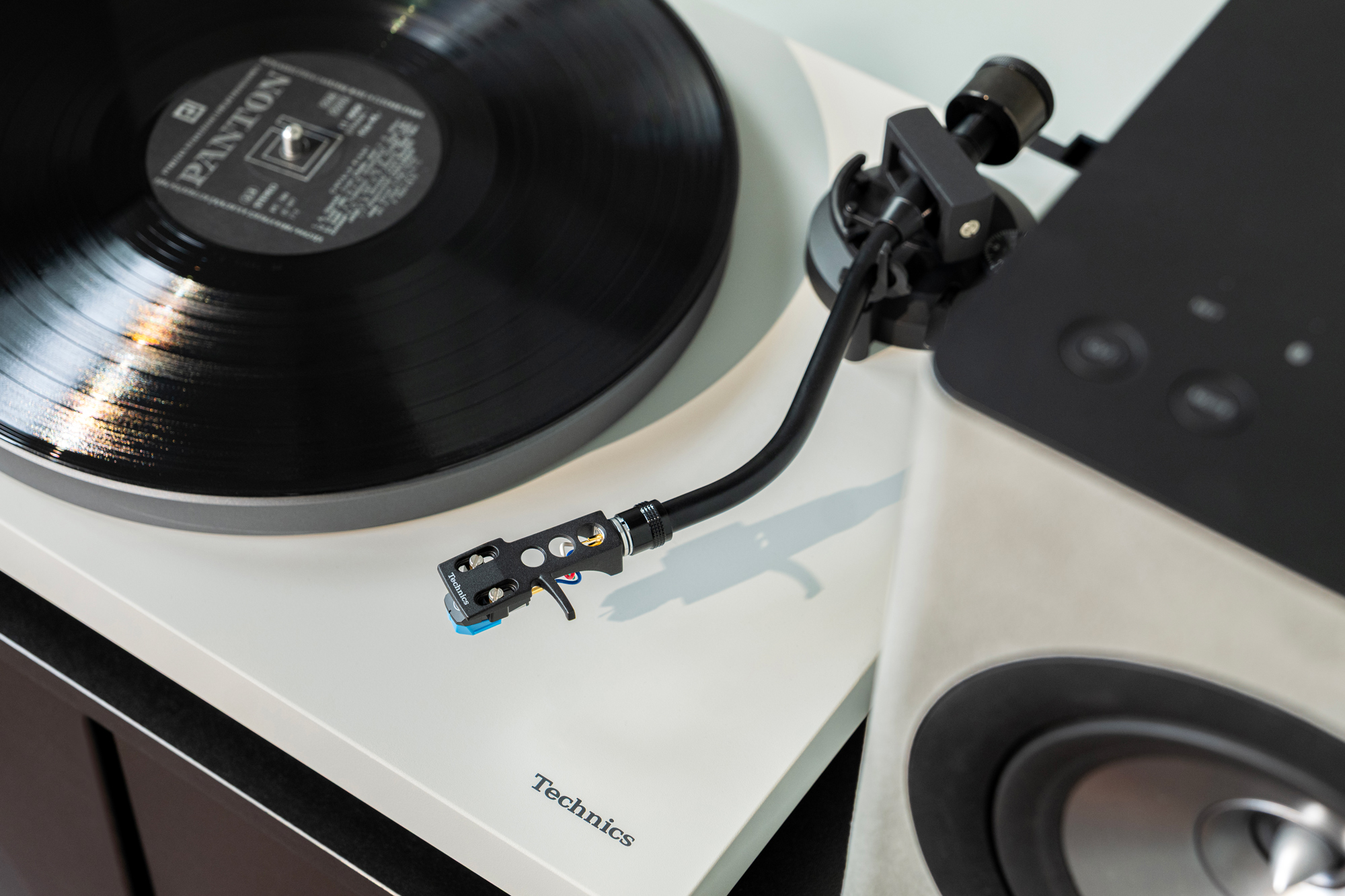 Gros plan sur les enceintes sans fil Technics CX700 et la platine Technics 40CBT. Les deux produits sont présentés en finition noire, dans un salon contemporain au design soigné. On distingue une main en train de manipuler les boutons situés sur le dessus de l’enceinte.
